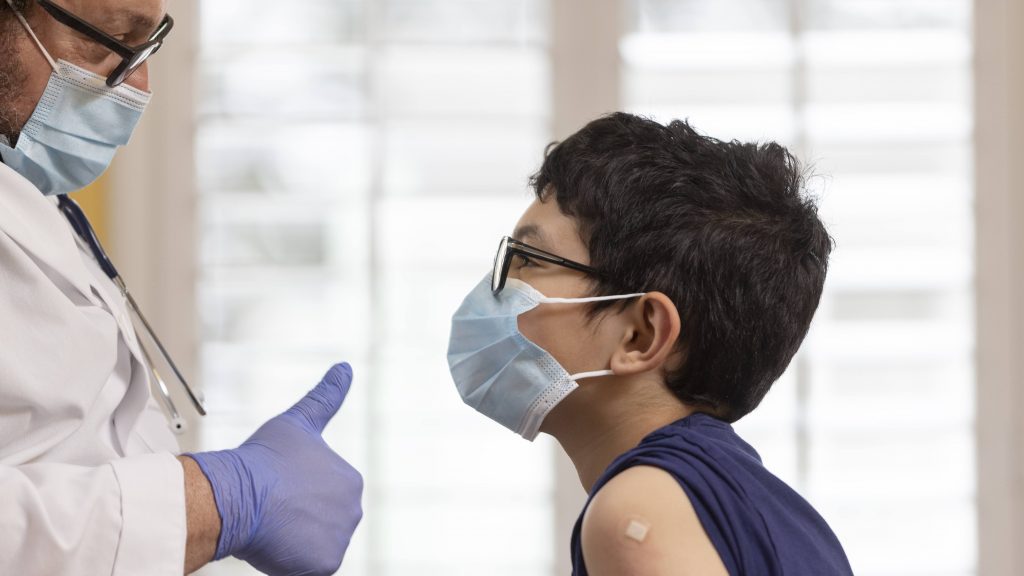 a young elementary school boy with glasses and wearing a mask with a bandaid on his arm after vaccine