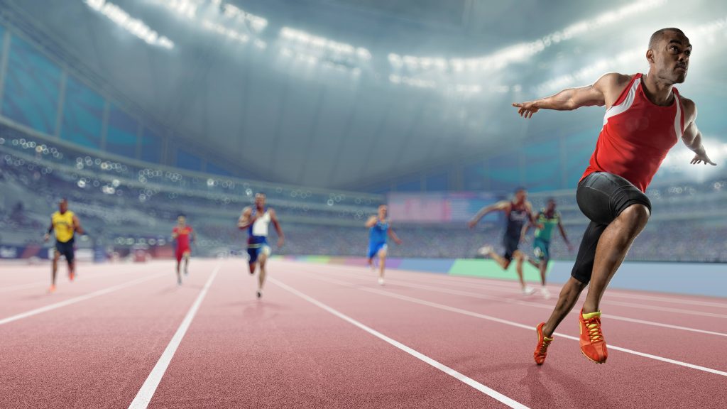 Wide angle, shallow depth of field image of a professional male track athlete sprinting over the finish line of a race with his arms outstretched in victory with composed expression. The athlete is competing in an indoor track sprint event in a generic arena, and finishes ahead of his competitors.