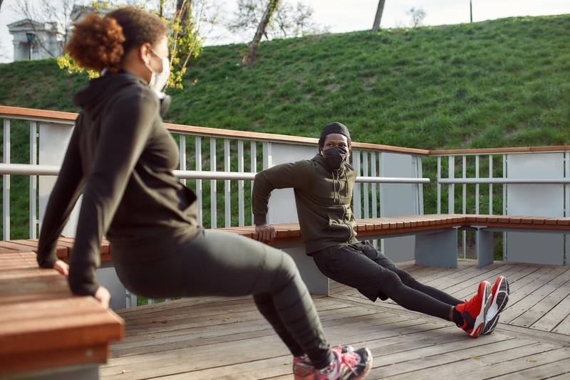 Masked couple working out in park outdoors perform tricep dips with help of built-in benches.