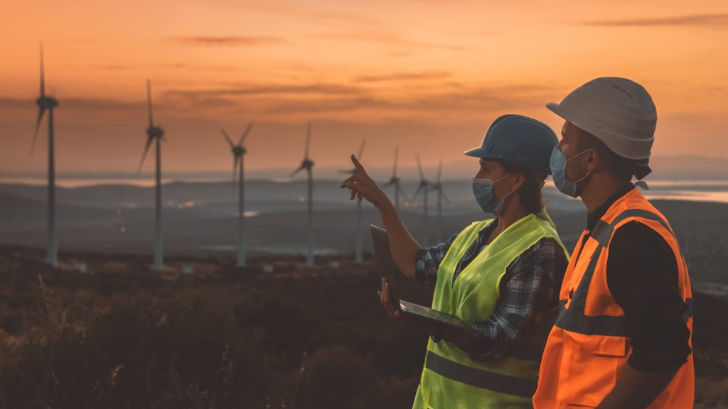 Young electrical engineer woman and businessman with face masks and safety vests working with laptop in wind turbine farm at sunset