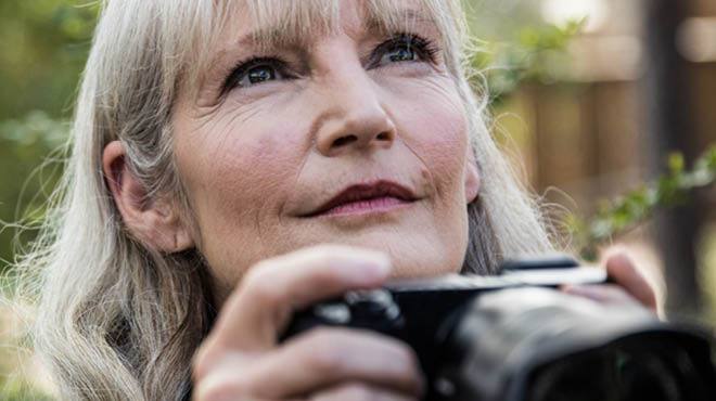 closeup of a smiling older woman outdoors, holding a camera and looking up