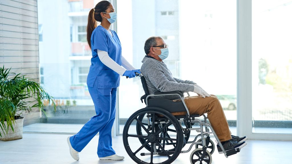 una joven enfermera enmascarada empujando a un anciano en silla de ruedas