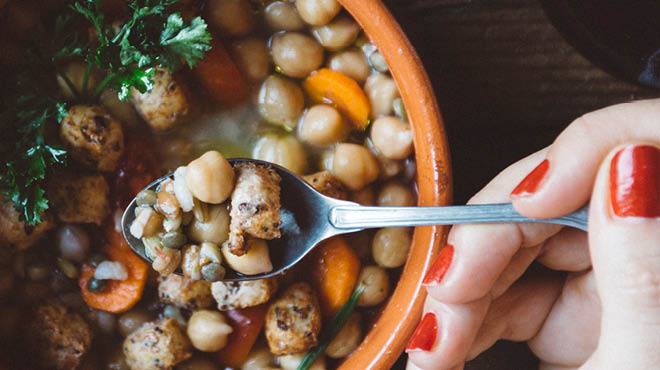 A hand holding a spoon over a bowl of soup with chickpeas