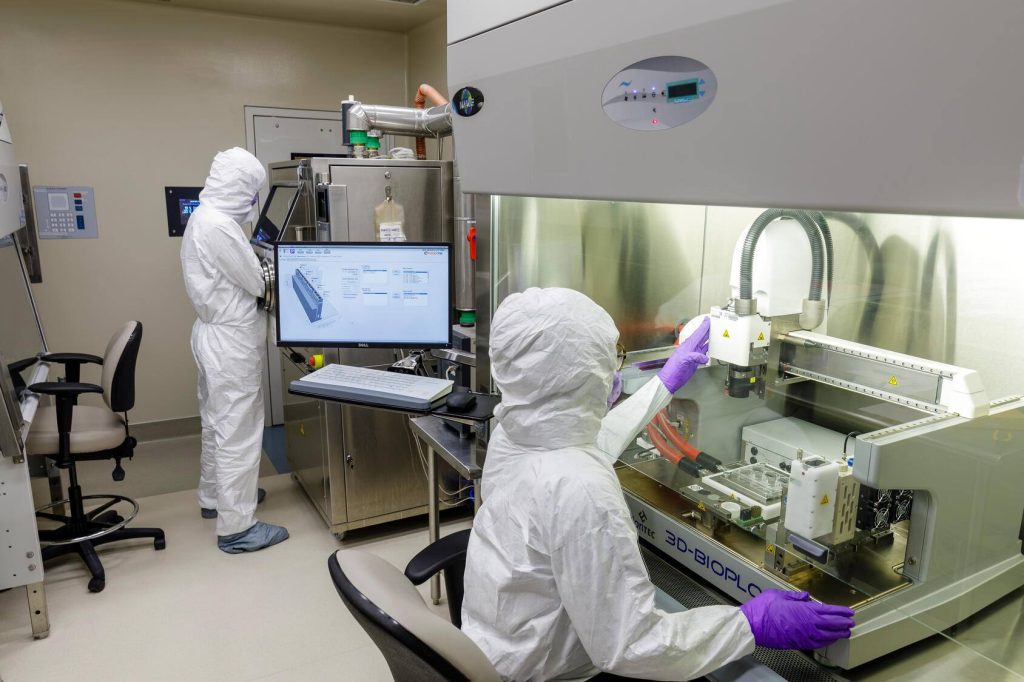 Two lab technicians wearing cleanroom suits operate bioprinters.