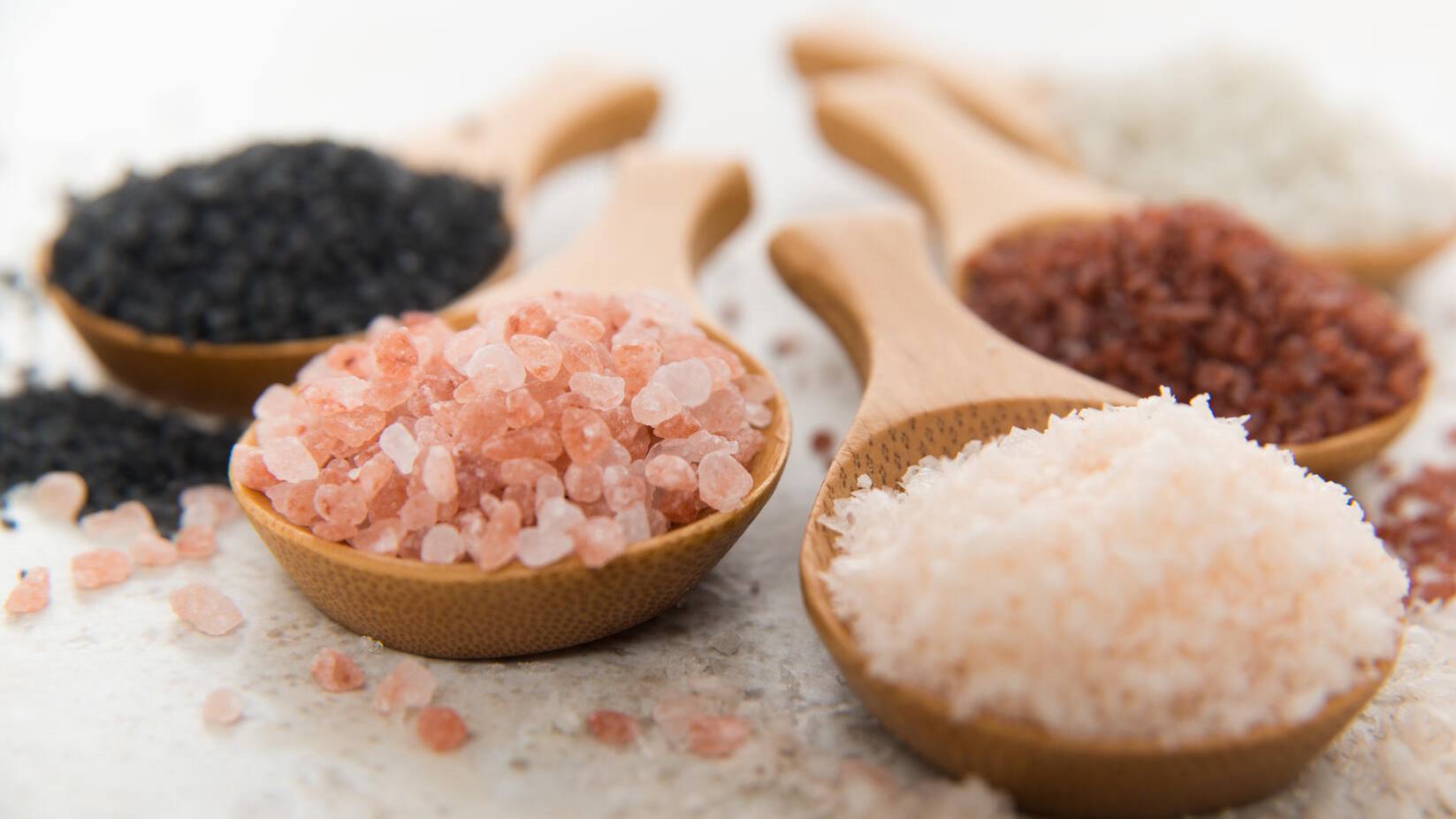 photograph of various types of sea salt in wooden spoons, black salt, pink salt, white salt, red salt,