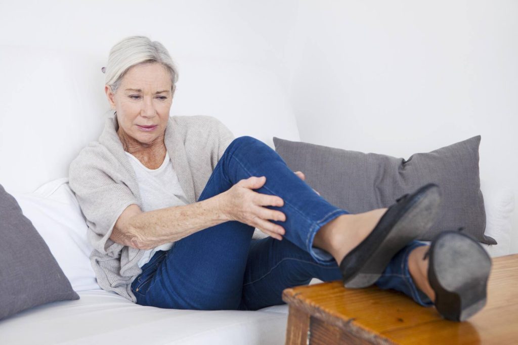 an older Caucasian woman sitting back on a couch with her feet up on an end table, holding her lower leg and looking concerned