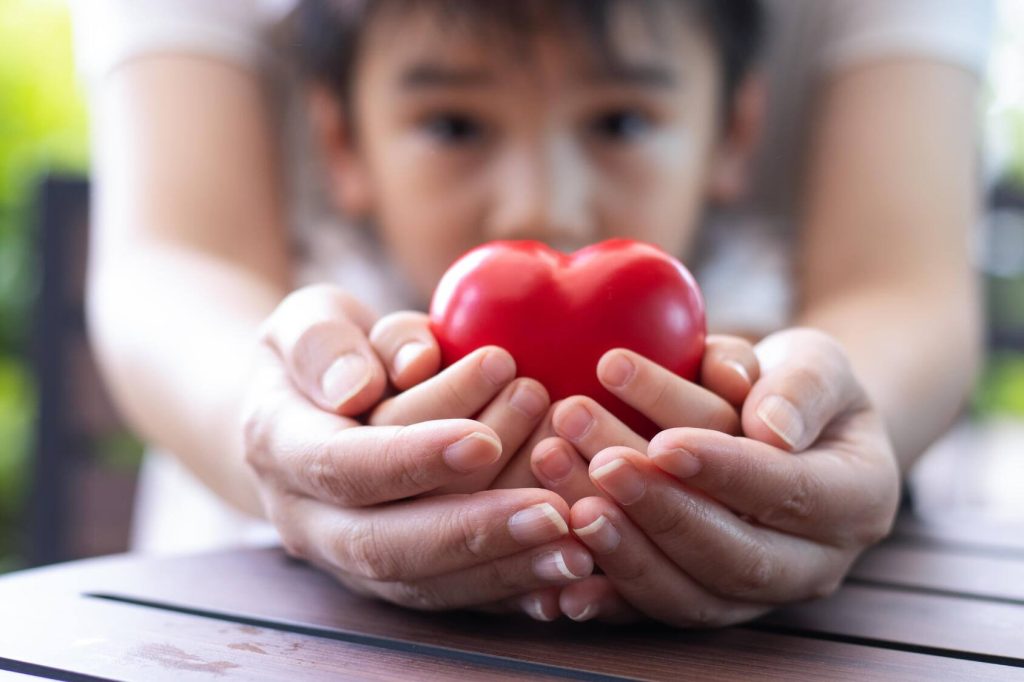 close-up of an adult's hands cradling a child's hands holding a red toy heart, with the child's face out of focus in the background