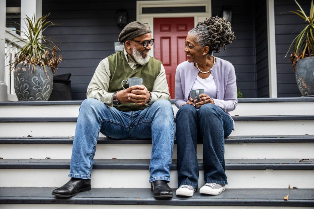 a Black man and woman sitting together on a front porch step, each holding a coffee cup, looking at one another and smiling