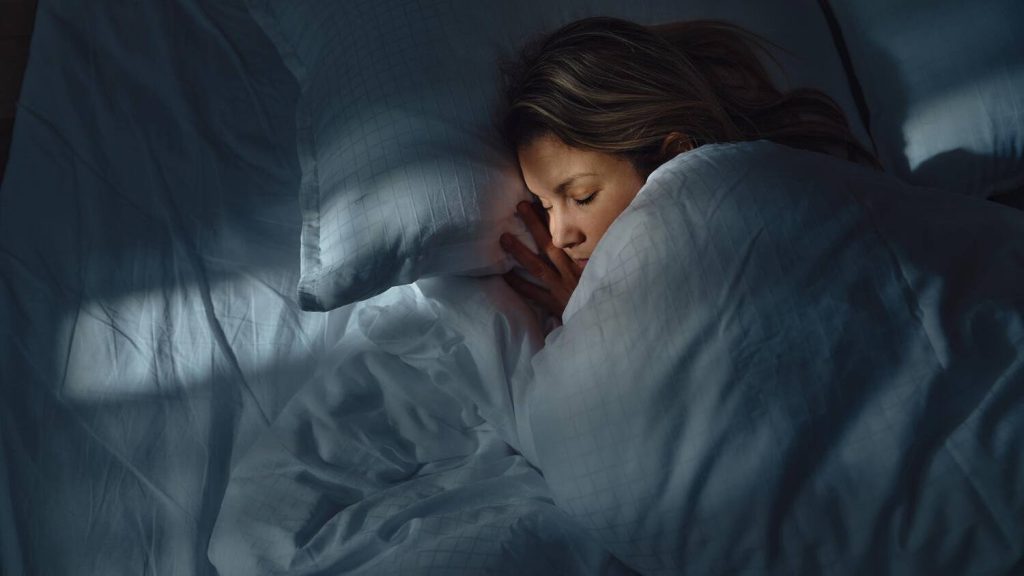 a white woman sleeping peacefully in bed in a darkened room, with a soft light falling across her face