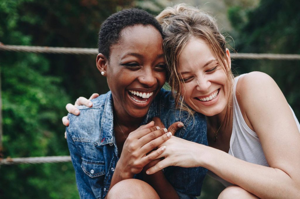 two young women, one Black and the other white, laughing and hugging outdoors