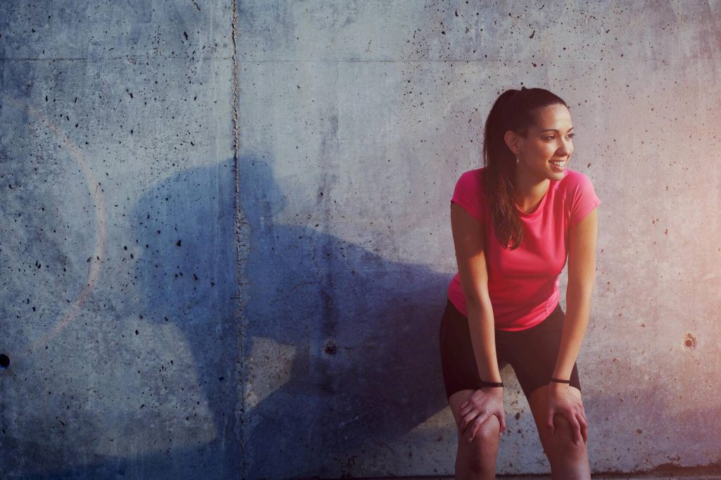 a smiling young woman in running clothes leaning against a concrete wall, with her shadow suggesting the sun is setting
