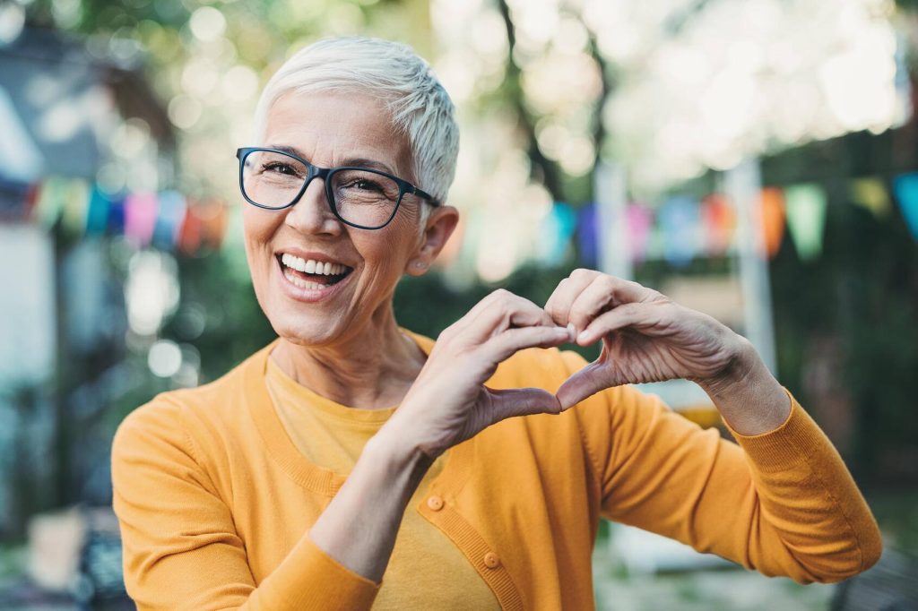 a smiling older white woman outdoors, wearing glasses and making a heart shape with her hands