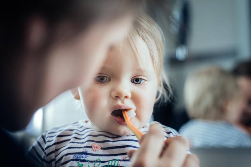 a close-up of an adult brushing the teeth of a very young white toddler with blond hair and blue eyes