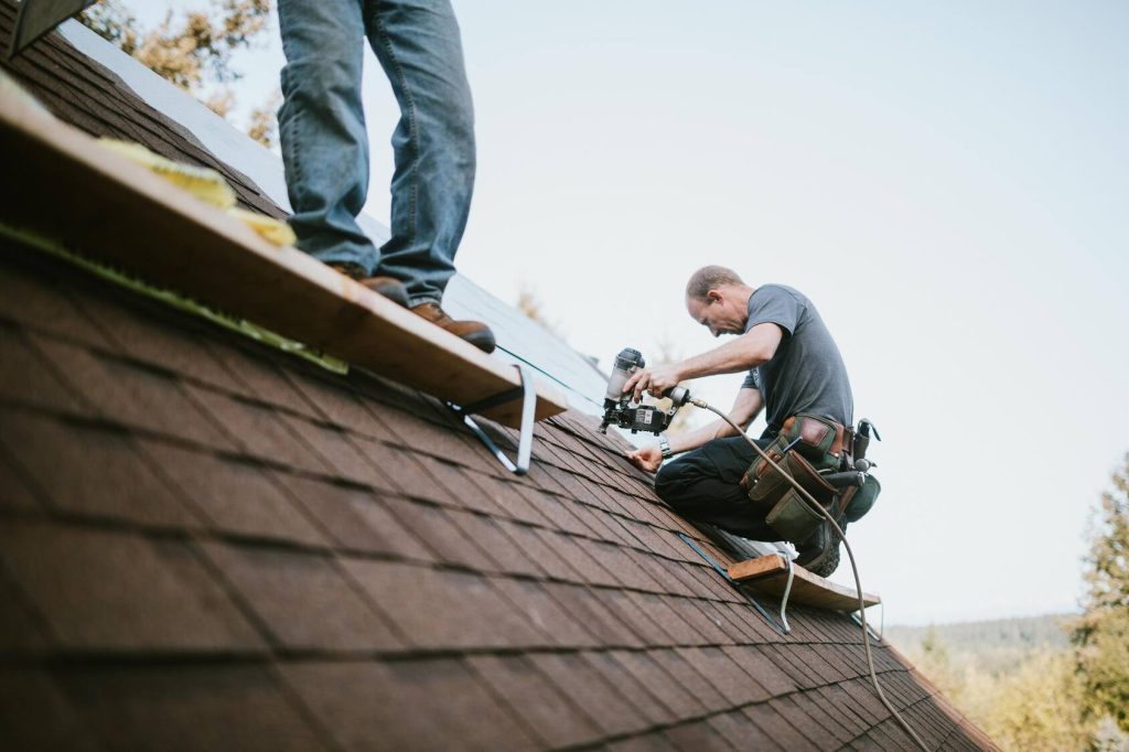 two workers replacing shingles on a roof, one holding a heavy pneumatic nail gun