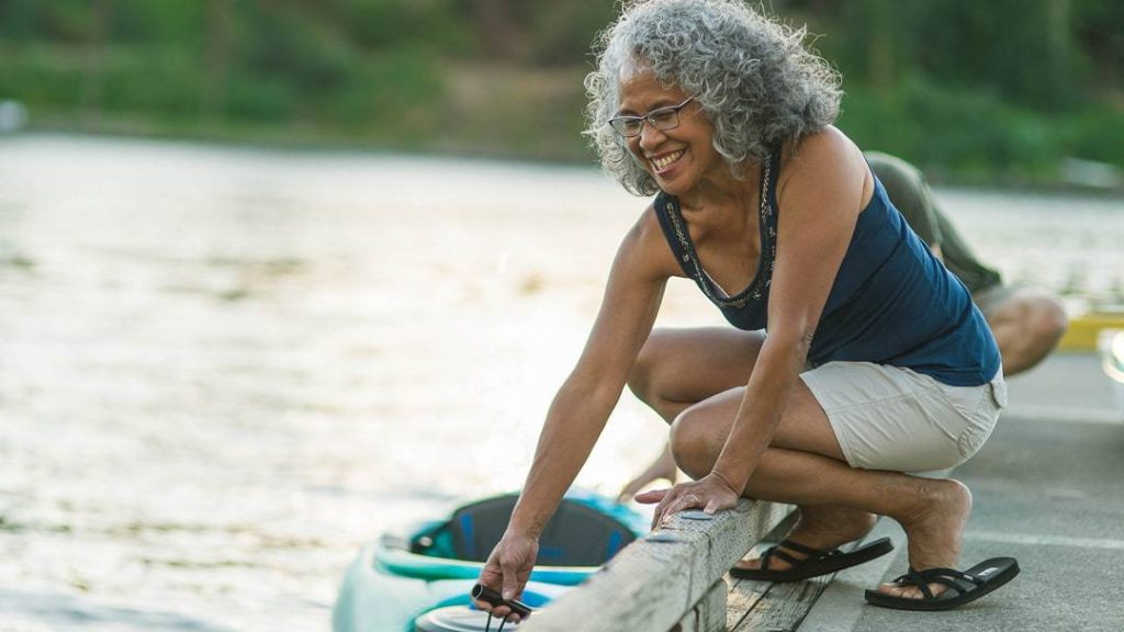 a smiling older Black woman crouching on a dock, pulling a kayak alongside
