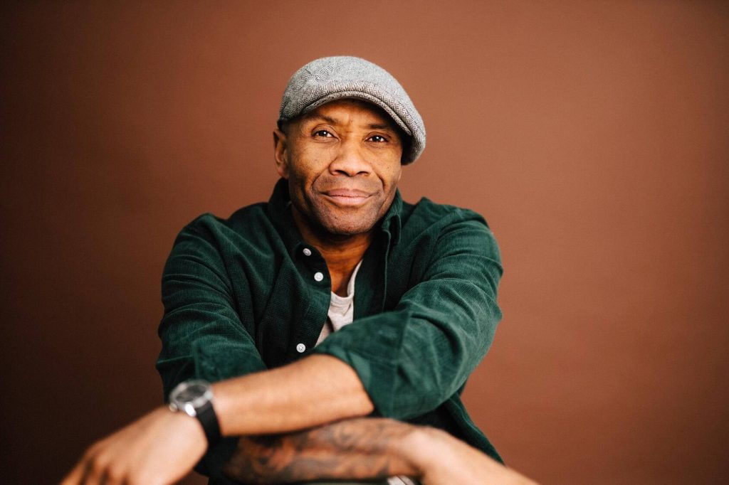 a portrait of a smiling middle-aged Black man wearing a grey herringbone cap and a dark green shirt