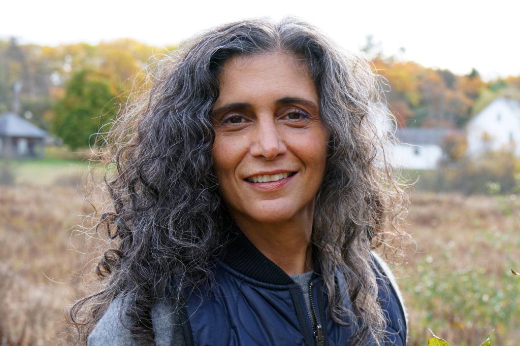 a closeup of a smiling middle-aged woman with long, curly, dark and greying hair, outside on a farm