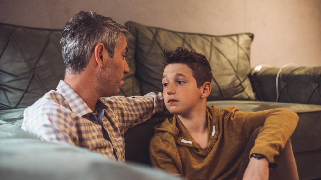 a white father and son having an emotional conversation seated on the floor in front of a couch