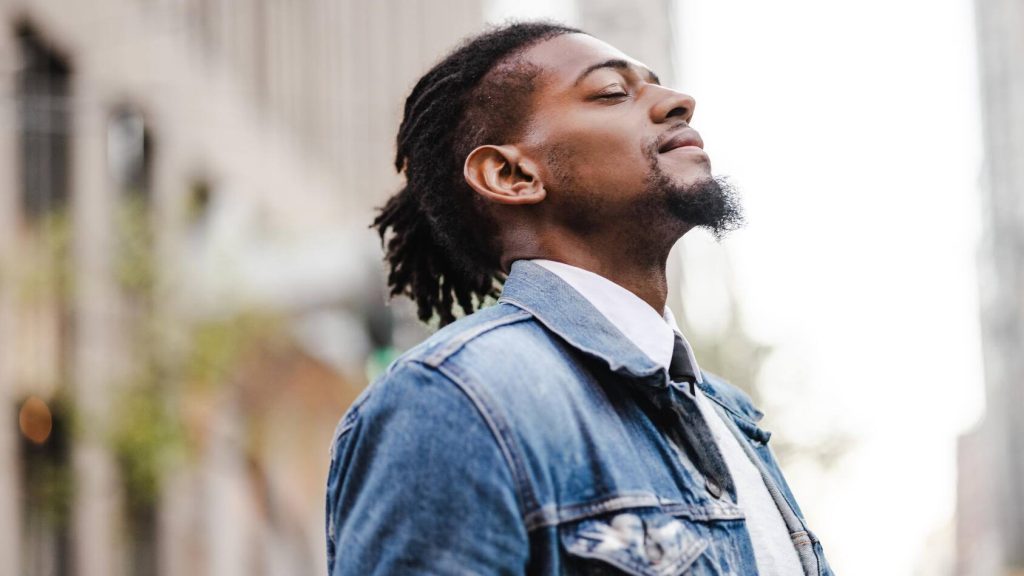 a young Black man standing on a city sidewalk, eyes closed and head back, appearing to take a deep breath