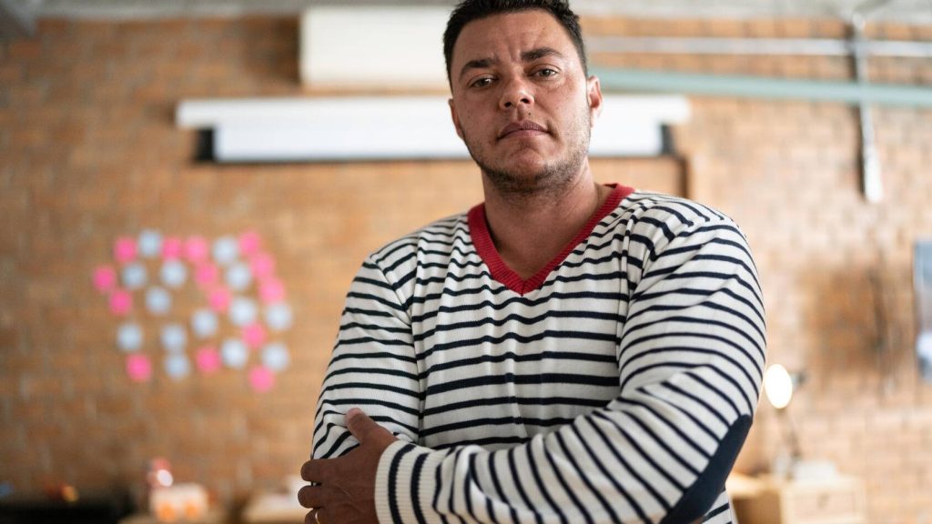 a young Latino man with a serious look on his face, standing with his arms crossed in a loft apartment or office