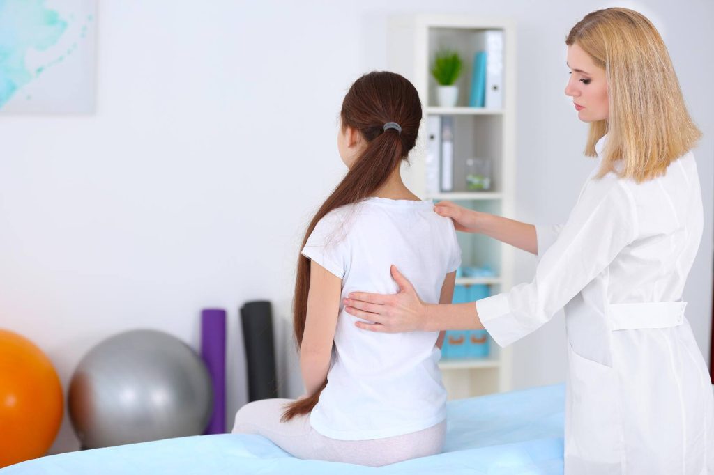 a young girl sitting on an examining table, with a healthcare professional examining her posture and back alignment