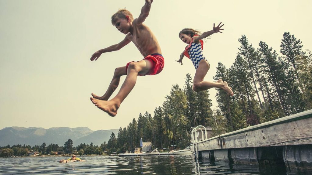 two young white children jumping off a dock into a lake, with mountains and evergreen trees in the background