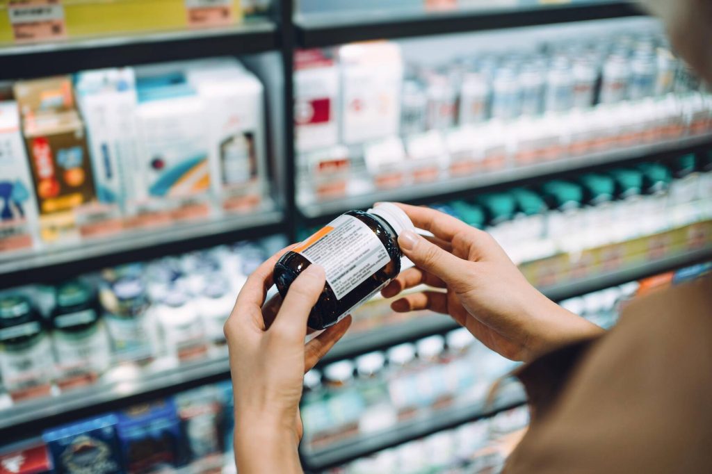 close-up of the hands of a white woman standing in a pharmacy aisle, reading the label on a bottle of vitamins or supplements, which may affect your blood pressure