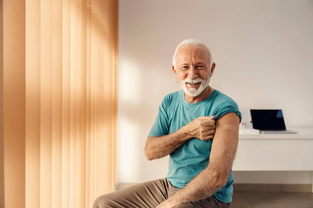 a smiling older white man in a doctor's office holding up one sleeve to show a bandaid where he'd been vaccinated