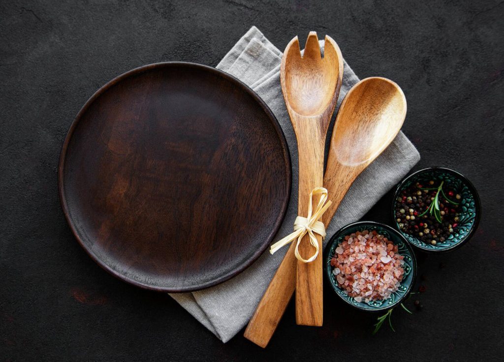 an empty wooden plate alongside a linen napkin, wooden serving utensils tied together with twine and small bowls of pink salt and whole peppercorns, all on a dark tabletop,  dietary

