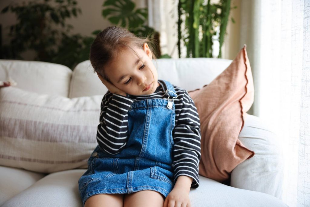 a young girl sitting on a couch with her head cocked to one side, looking sad and holding her hand up to her ear