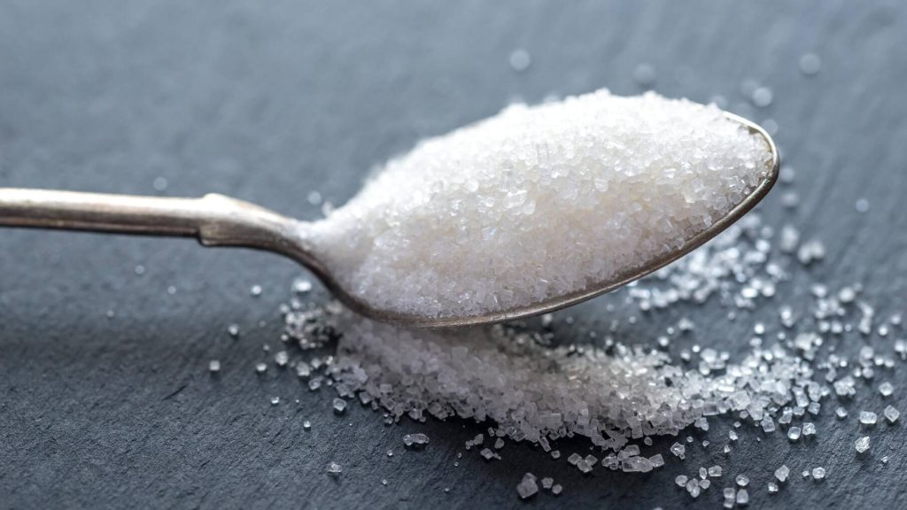 a silver teaspoon overflowing with white granulated sugar, spilling out onto the gray counter surface