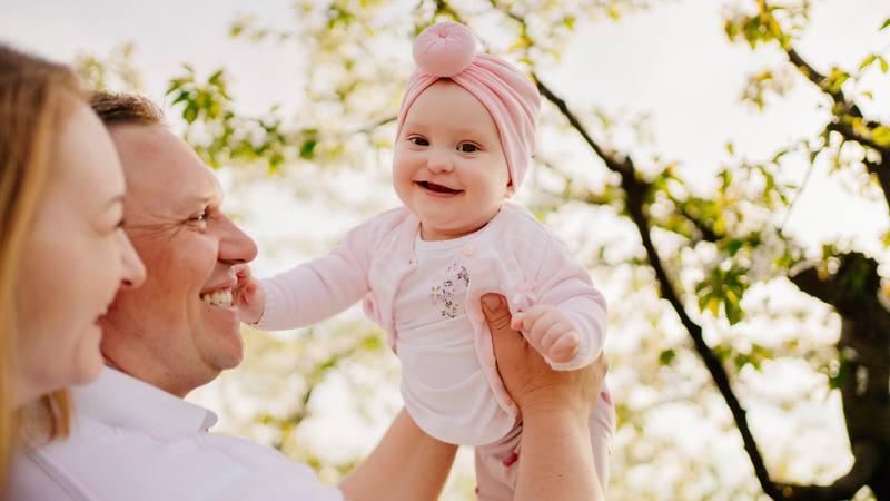 A man holds up a baby, who has had surgery for a cleft lip, next to a woman, as they all smile together near a green tree.