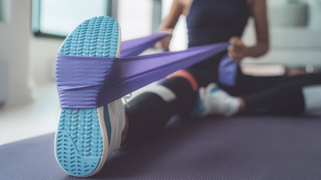 Resistance band exercise at home. Woman doing pilates workout using elastic strap pulling with arms for shoulder training on yoga mat indoors.