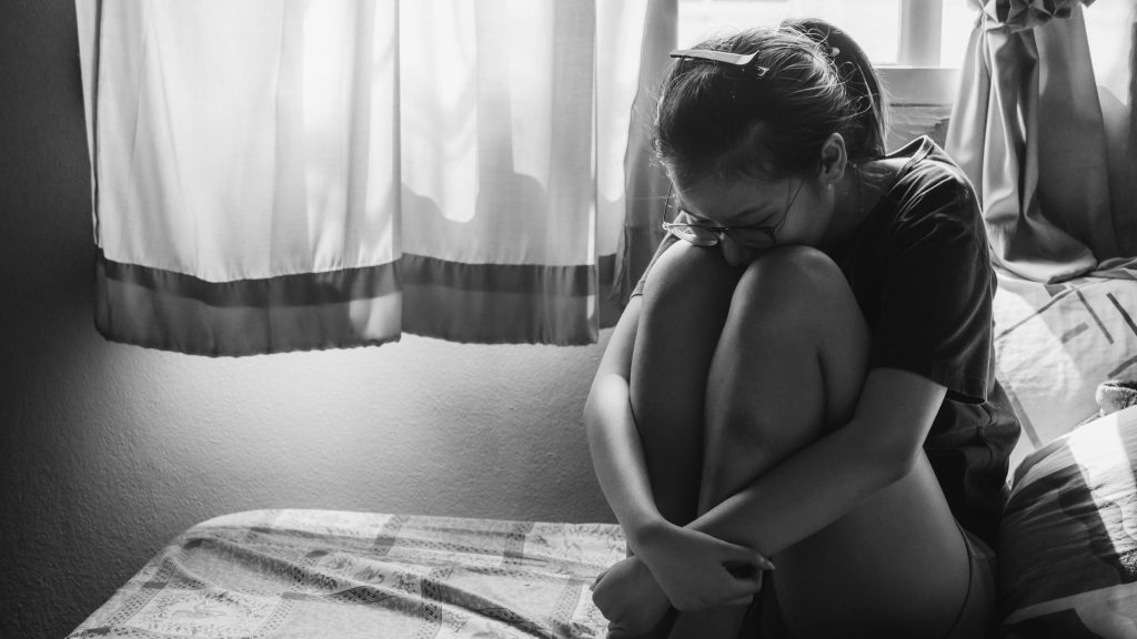 Black and white image of a teen girl, looking dpressed, sitting on a bed in front of a window - Getty /Boy_Anupong