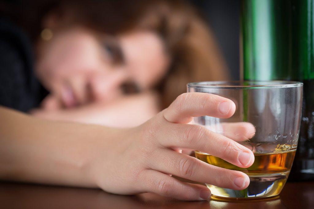 woman holding a glass of whisky, rum, or liquor while resting her head on the bar, focus on the glass of alcohol with her face blurred. Depiction of addiction, alcoholism, depression.