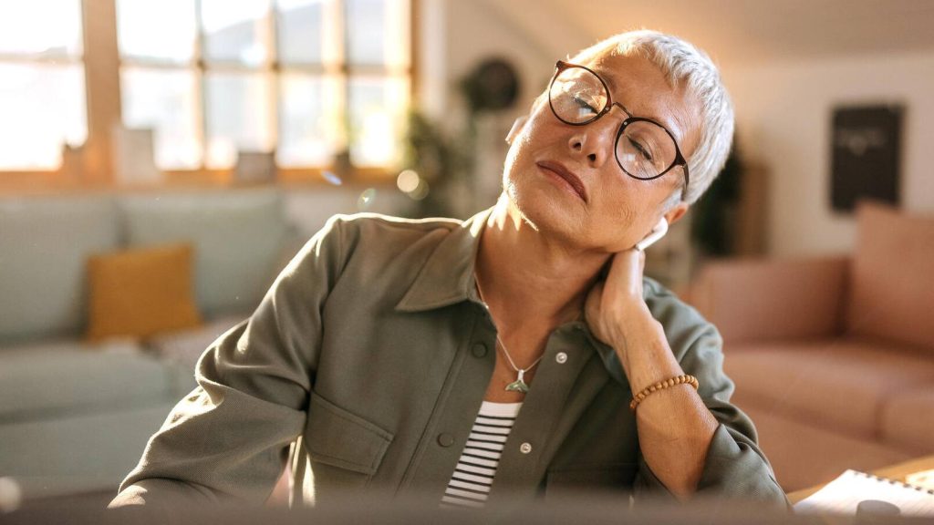 Middle aged woman, short white hair, rubs neck, serious, somber, tired