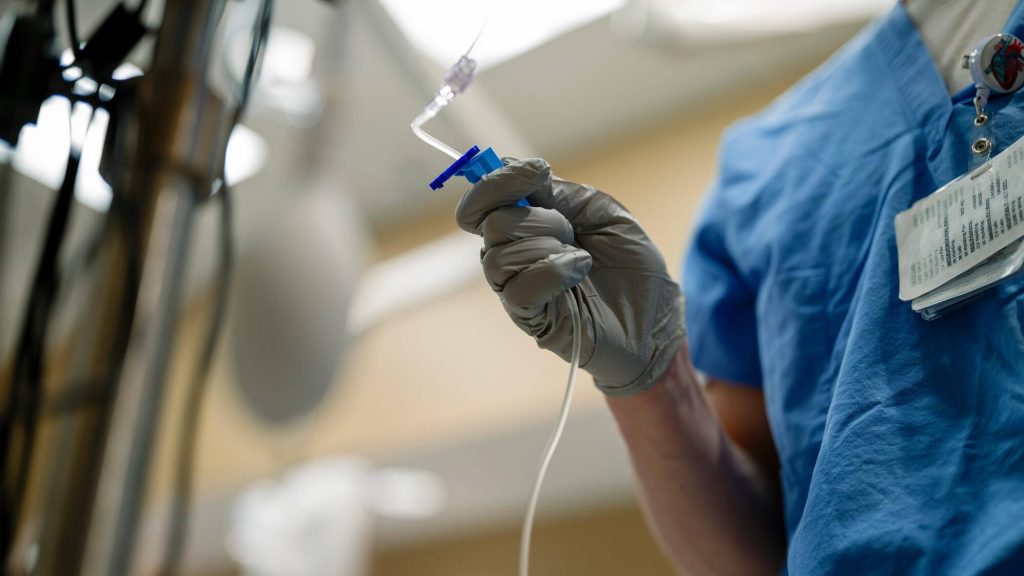 A gloved hand adjusts the roller clamp on an intravenous (IV) drip line