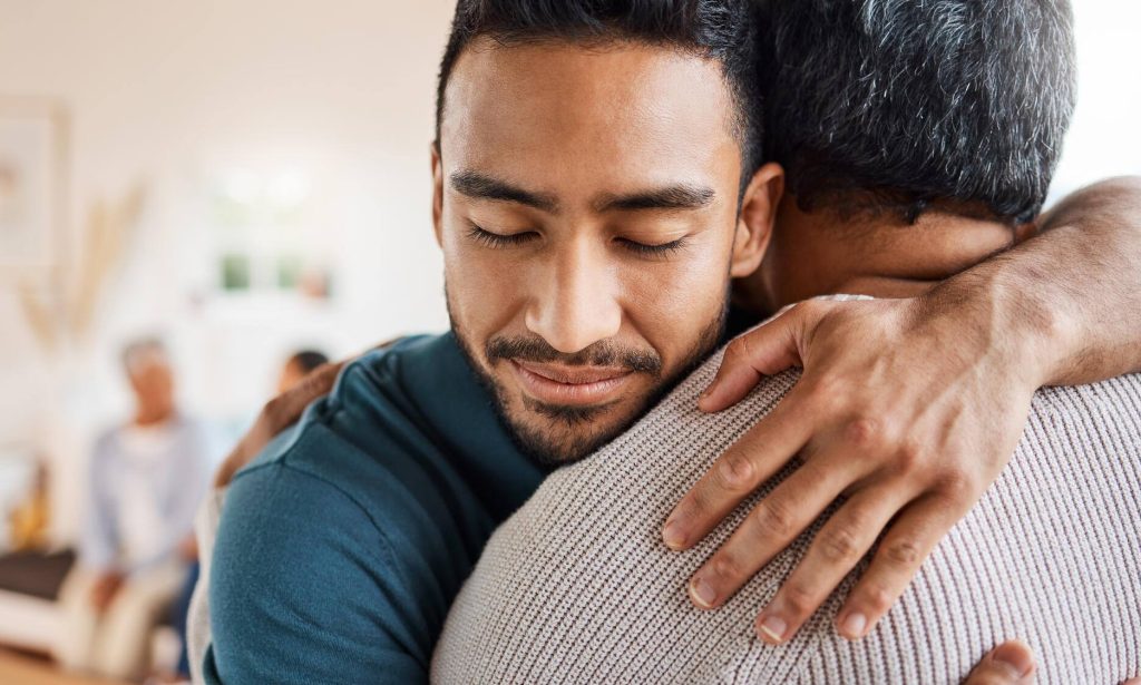 A man smiles with his eyes closed as he hugs an older man