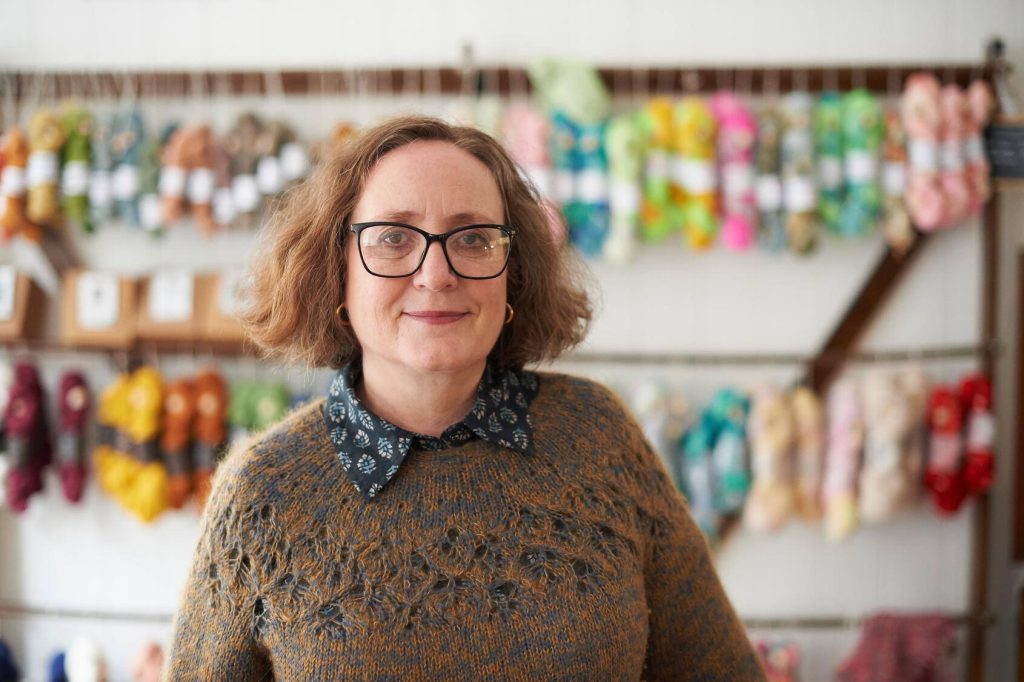 Portrait if a middle-aged woman yarn store owner standing in front of a rack of yarn