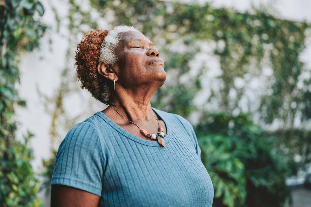 A woman with closed eyes tips her head back and takes a deep breath while standing outdoors.