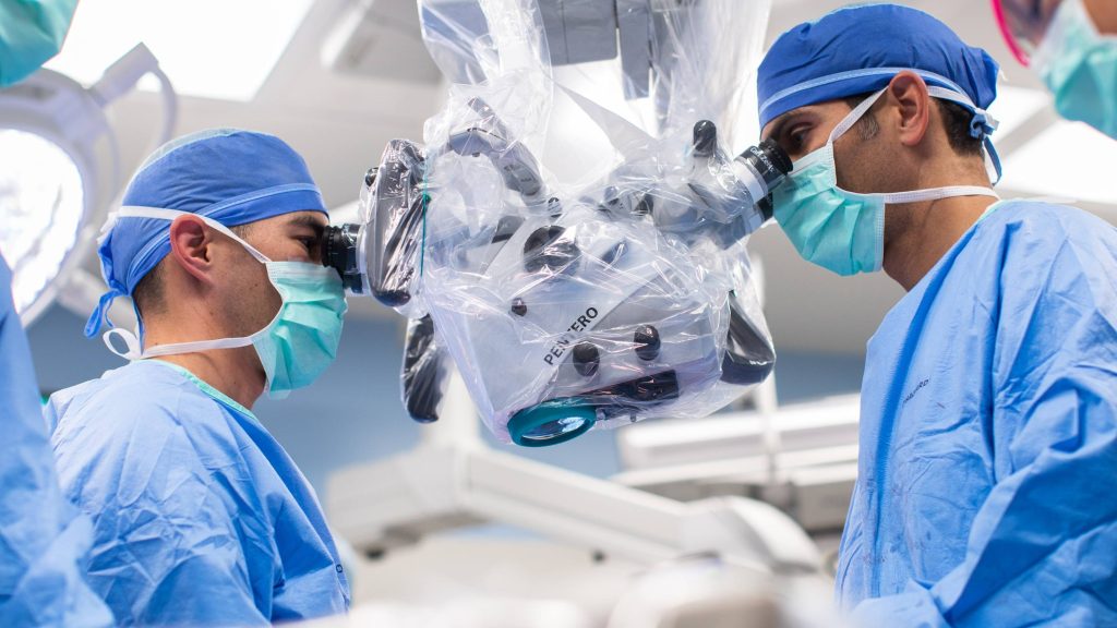 Dr. Kevin Arce and Dr. Kyle Ettinger from Division of Oral and Maxillofacial Surgery in the operating room (OR). Both doctors are looking through a microscope while wearing scrubs and personal protective equipment (PPE).