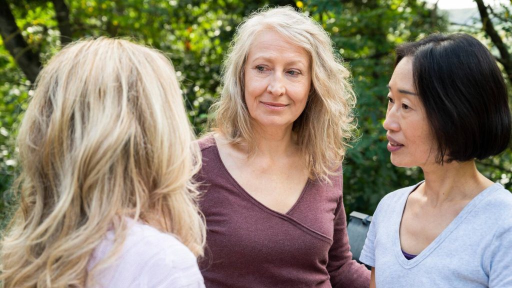 Two women listen carefully to their friend speak while standing outside.