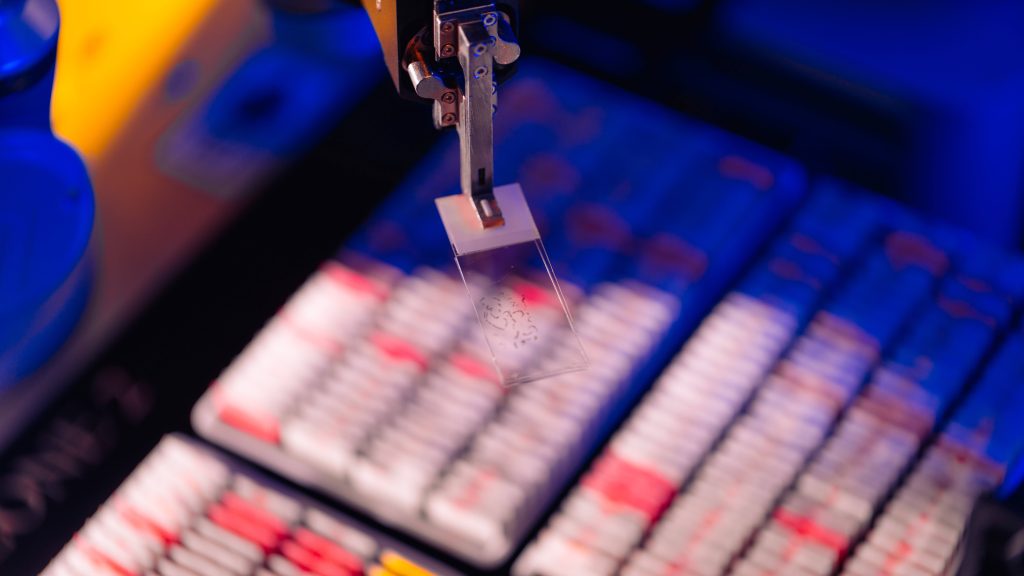 Close-up of a robotic arm holding a glass microscope slide above rows of blurred laboratory sample trays under blue and pink lighting, suggesting an automated digital pathology setting.