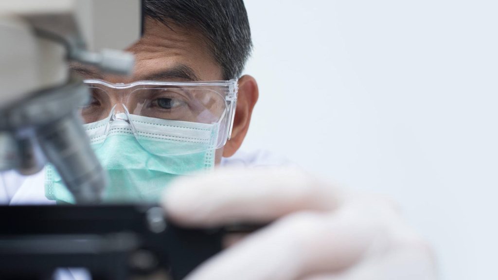 A doctor or scientist wearing gloves, protective face mask, and safety glasses, looking at a chemical sample through a microscope.