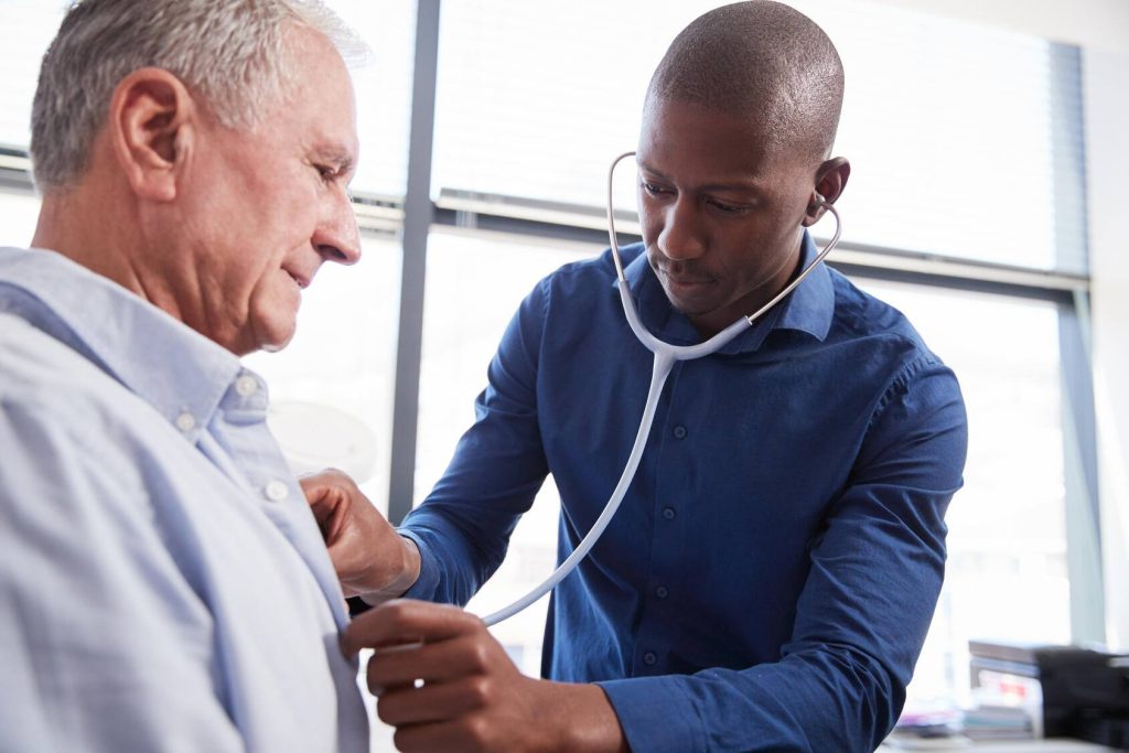 A close up of a physician leaning over and using a stethoscope to listen to the chest of a seated patient. The physician is conducting this exam in his office.