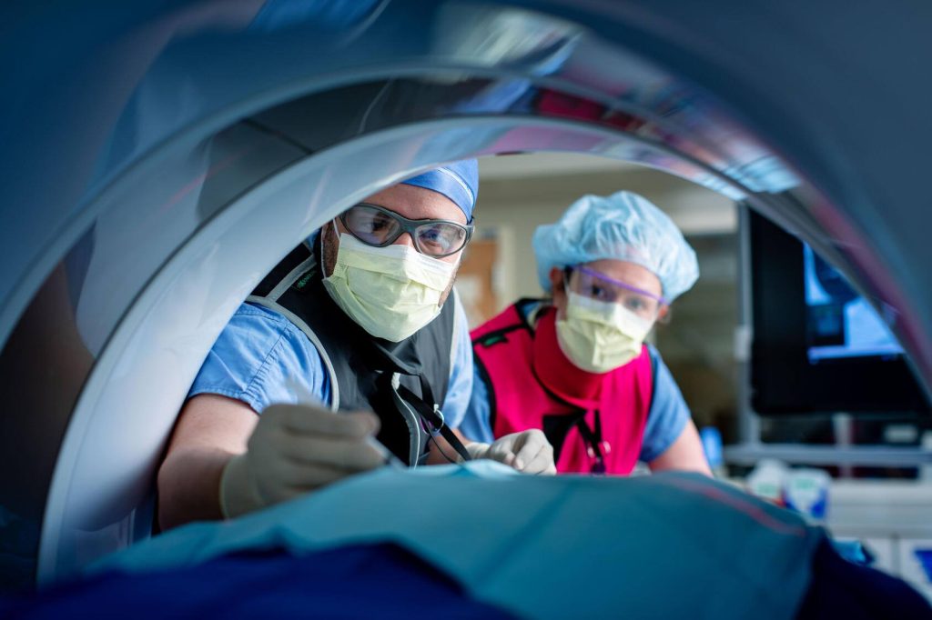 Two healthcare professionals wearing protective eyewear and lead aprons lean over a patient positioned inside a large medical imaging scanner during an image-guided procedure.