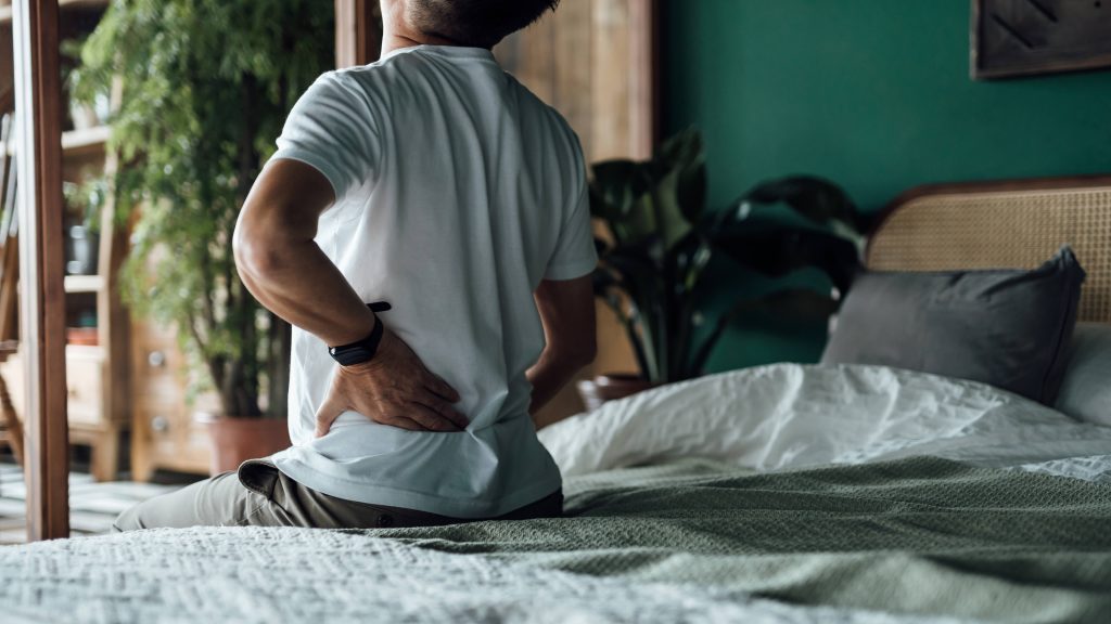 Vended Stock - Getty; A rear view of man sitting on his bed and massaging his lower back muscles to relieve the pain.