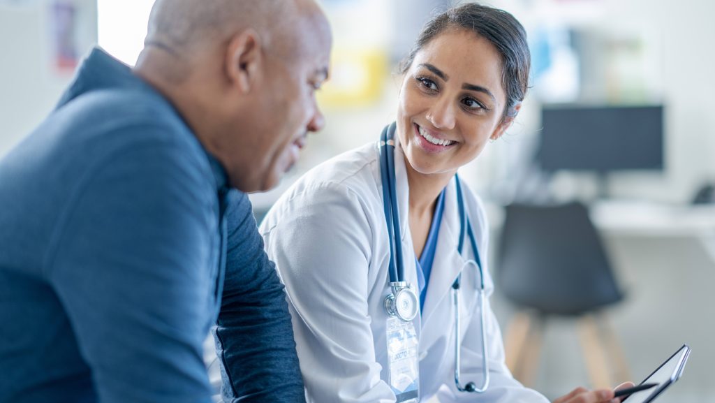 A doctor of sits casually beside a patient in an exam room, with a digital tablet in hand