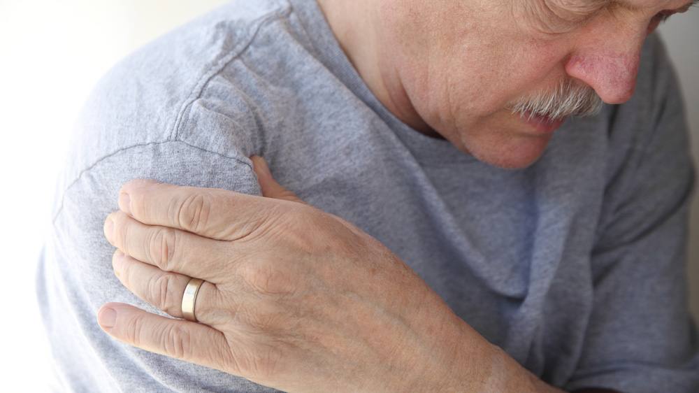 Closeup of man with head bent forward, holding his hand to his shoulder as if in pain or discomfort.