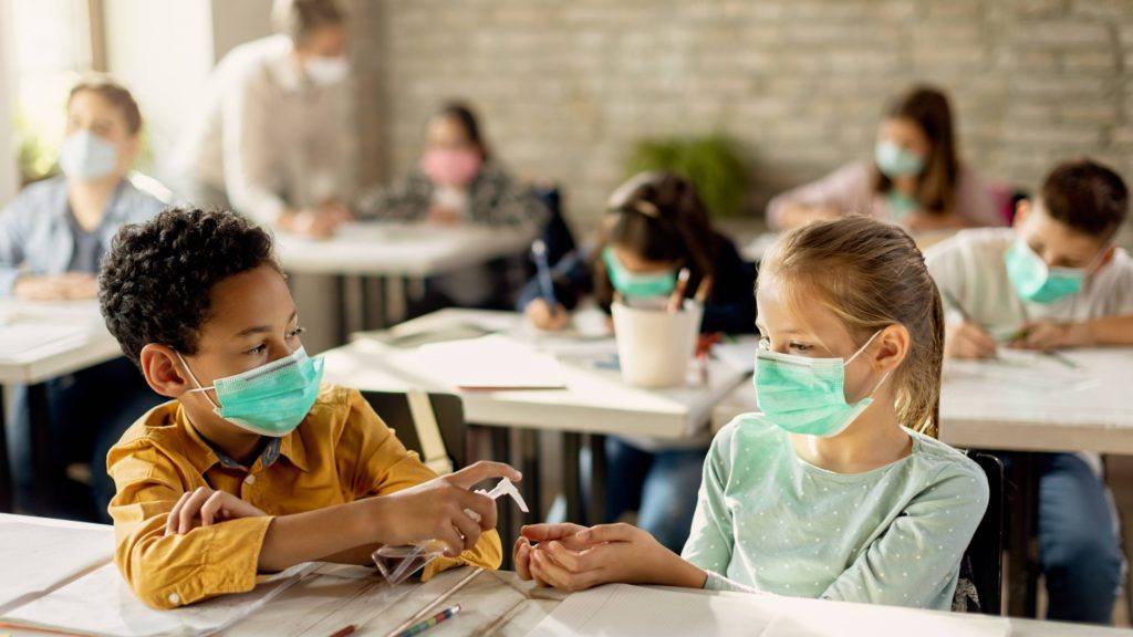 两个年幼的孩子，一个黑人男孩和一个白人女孩，在学校的教室里戴着口罩，坐在桌子前共用洗手液